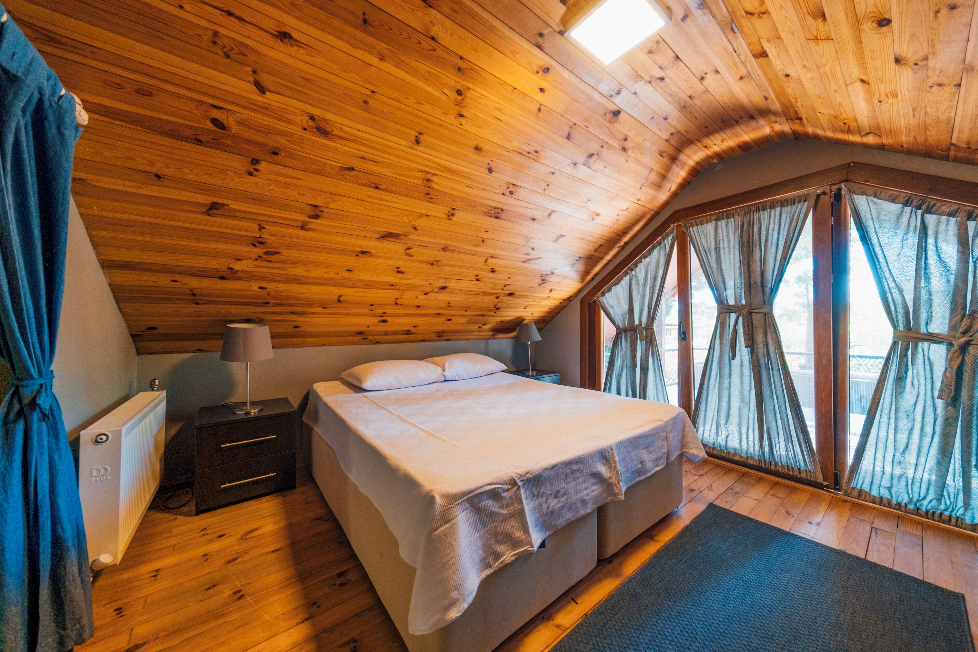 Un dormitorio con techo de madera, cama con ropa blanca, mesita de noche, lámpara y ventana con cortinas.