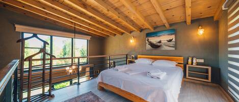 A bedroom with a wooden ceiling, a large bed, a nightstand, a wall-mounted light, and a window with a view of greenery.