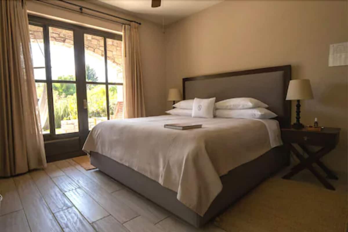 Une chambre avec un grand lit, une fenêtre avec des rideaux, une table de chevet avec une lampe, et une vue sur la verdure à l’extérieur.