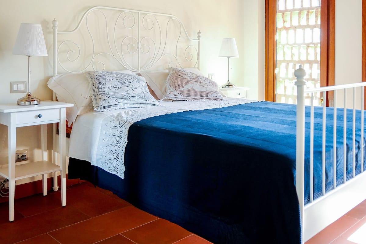 A bedroom with a white metal bed frame, a blue bedspread, two bedside tables with lamps, a wooden door, and a ceiling with exposed beams.