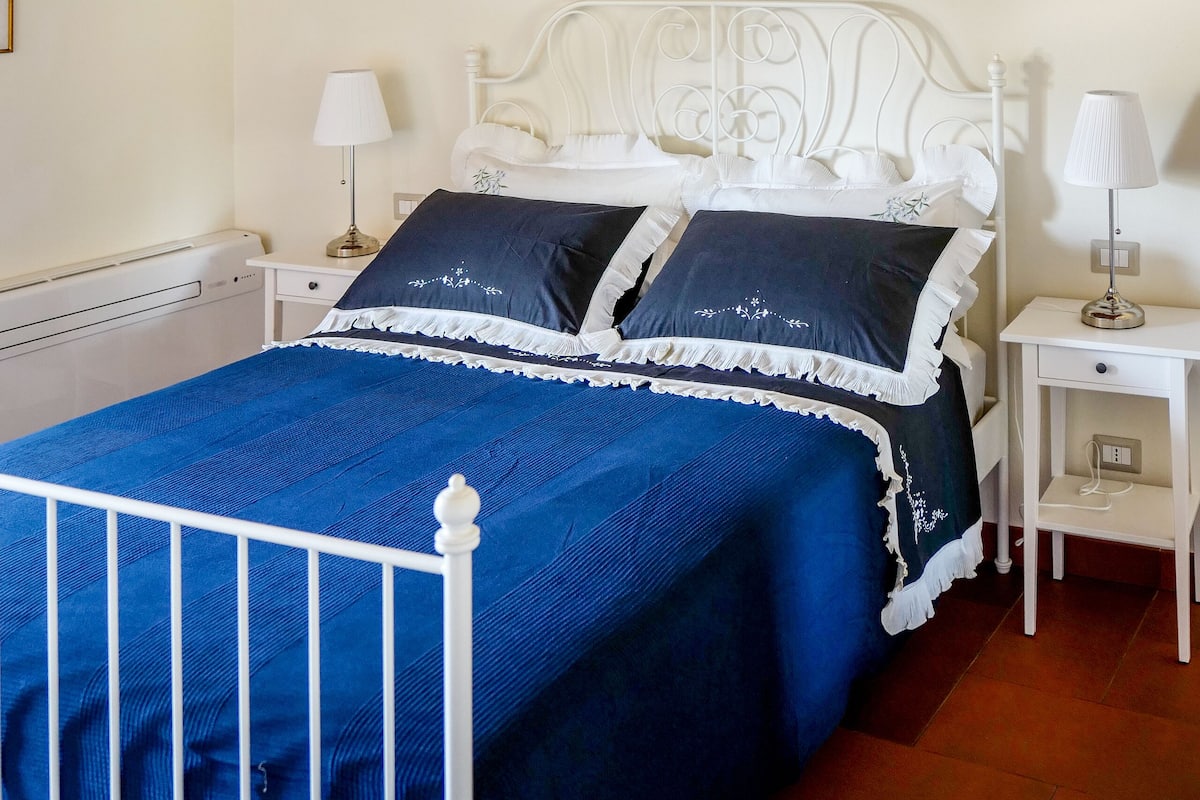 A bedroom with a white metal bed frame, a blue bedspread, white nightstands, a white lamp, a painting on the wall, and a wooden ceiling.