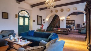 Ein geräumiges Wohnzimmer mit einem großen blauen Sofa, einem Holztisch und einer Kronleuchter. Eine Treppe führt zu einer oberen Ebene, und es gibt einen Essbereich mit Tisch für vier Personen.