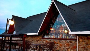 A stone building with a sloped roof, outdoor seating with metal chairs, and a bar area with bottles.