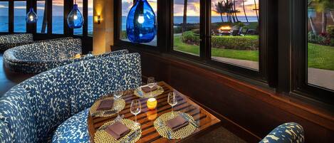 Un restaurante con asientos de estampado azul, una mesa de madera dispuesta con vasos y cubiertos, y vista al océano y palmeras al atardecer.