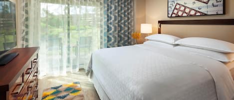 A hotel room with a bed, a desk, a chair, a lamp, and a window with sheer curtains.