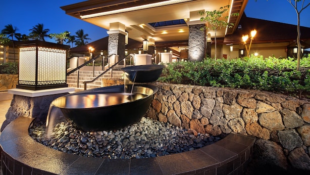 Un resort con un muro de piedra, una gran fuente de agua en un recipiente negro y una escalera que conduce a la entrada.