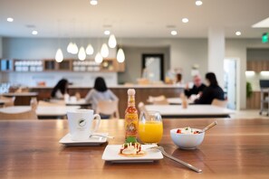 Une table en bois dressée pour le petit-déjeuner, avec une assiette de nourriture, un verre de jus d’orange et une bouteille de sauce piquante.