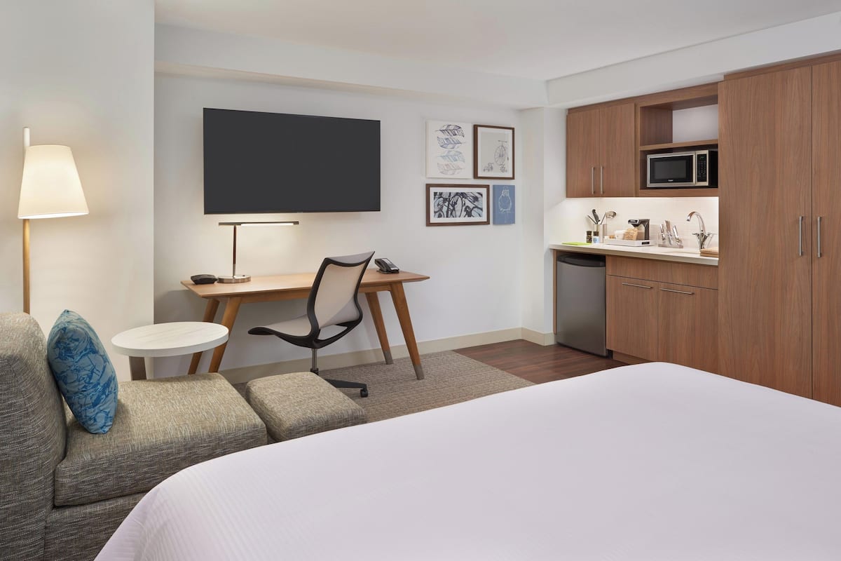 A hotel room with a bed, a desk, a chair, a small table, a lamp, a microwave, and a dishwasher.