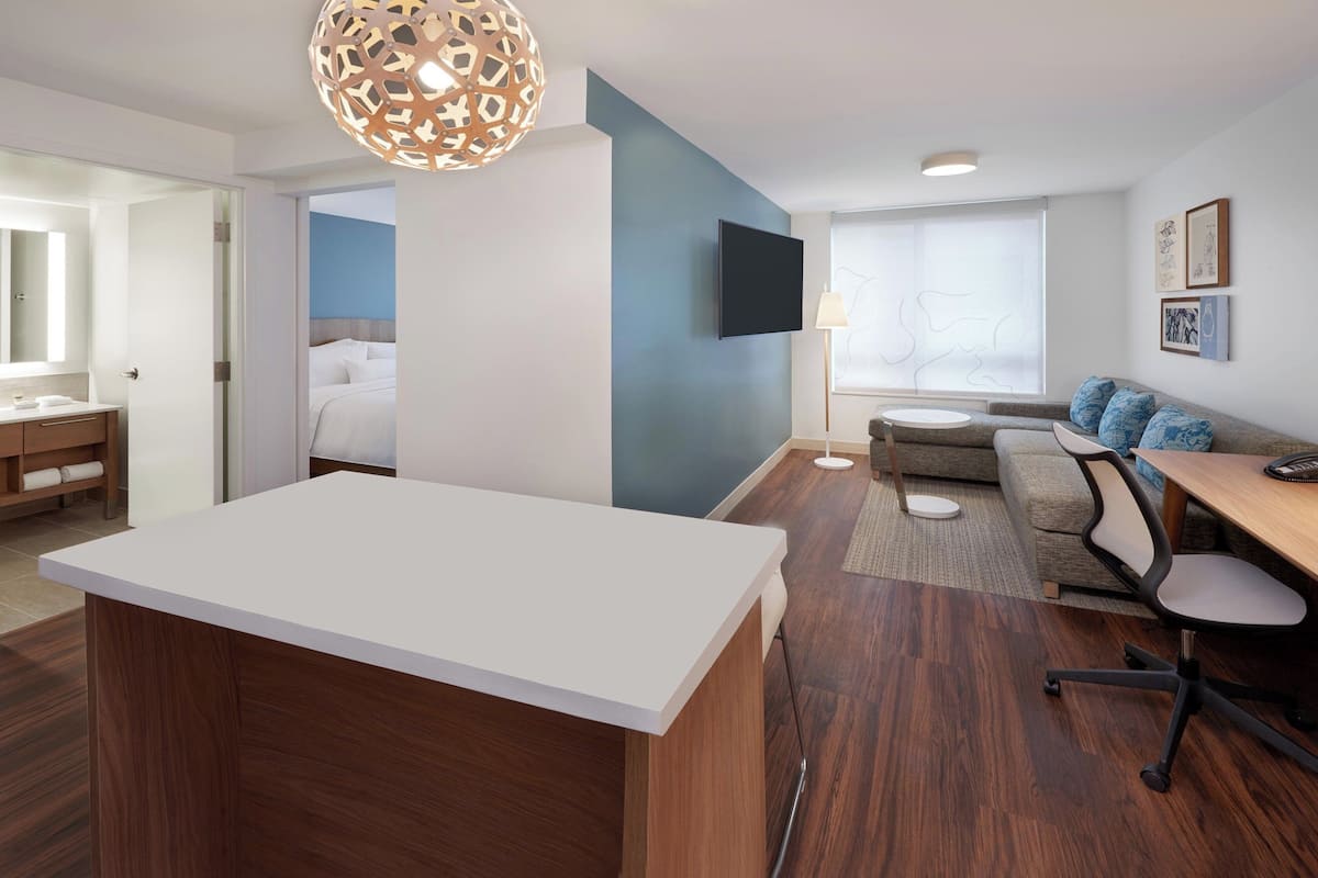 A modern hotel room with a wooden desk, a sofa with blue cushions, a flat-screen TV, and a large window.