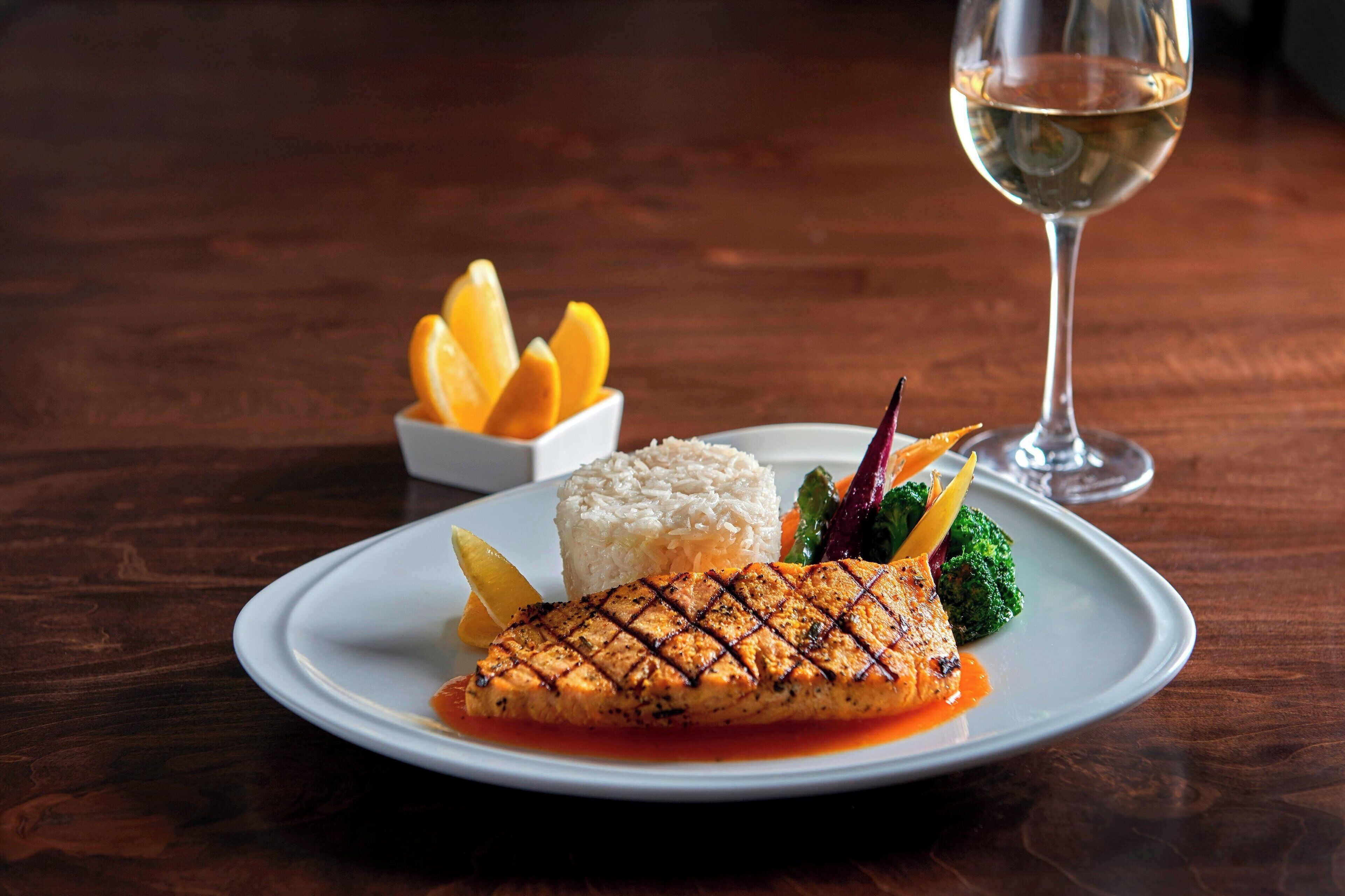 A plate of grilled salmon with rice and vegetables, accompanied by a glass of white wine on a wooden table.
