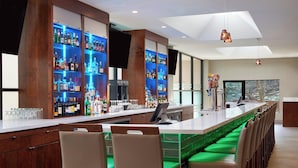 Un área de bar moderna con una barra larga, múltiples estantes con botellas de licores y taburetes iluminados con luz verde.