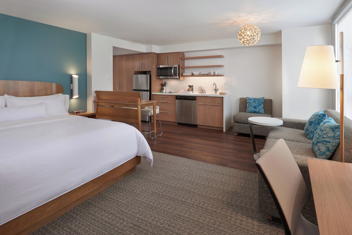 A modern hotel room with a large bed, a kitchenette, a dining area, and a seating space.