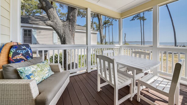 A covered porch with a white table and chairs, a wicker sofa with colorful cushions, and a view of the beach and trees.