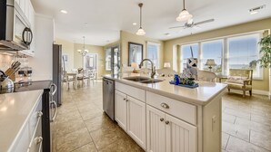 A spacious kitchen with white cabinetry, a large island, and a dishwasher. There is a dining area with a table and chairs, and a ceiling fan.