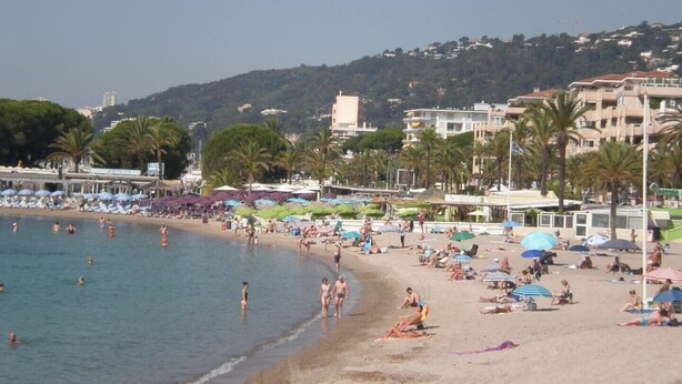 A beach with people sunbathing and swimming, umbrellas, and buildings in the background.