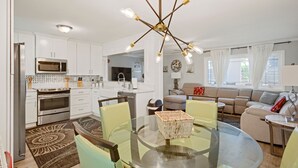Eine moderne Küche mit weißen Schränken, einem Edelstahl-Kühlschrank und einer Mikrowelle. Es gibt einen Essbereich mit einem Glastisch und Stühlen, sowie ein Wohnzimmer mit einem beigen Sofa und einer Lampe.