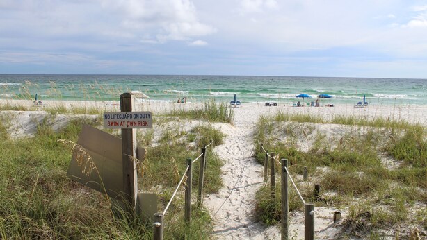 Ein Strandweg, der zum Meer führt, mit einem Schild, das daraufhinweist, dass keine Badeaufsicht vorhanden ist und Schwimmen auf eigene Gefahr.