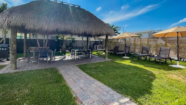 A thatched-roof outdoor seating area with tables and chairs, surrounded by a well-maintained lawn and a brick pathway. - Greniers (Hollywood)