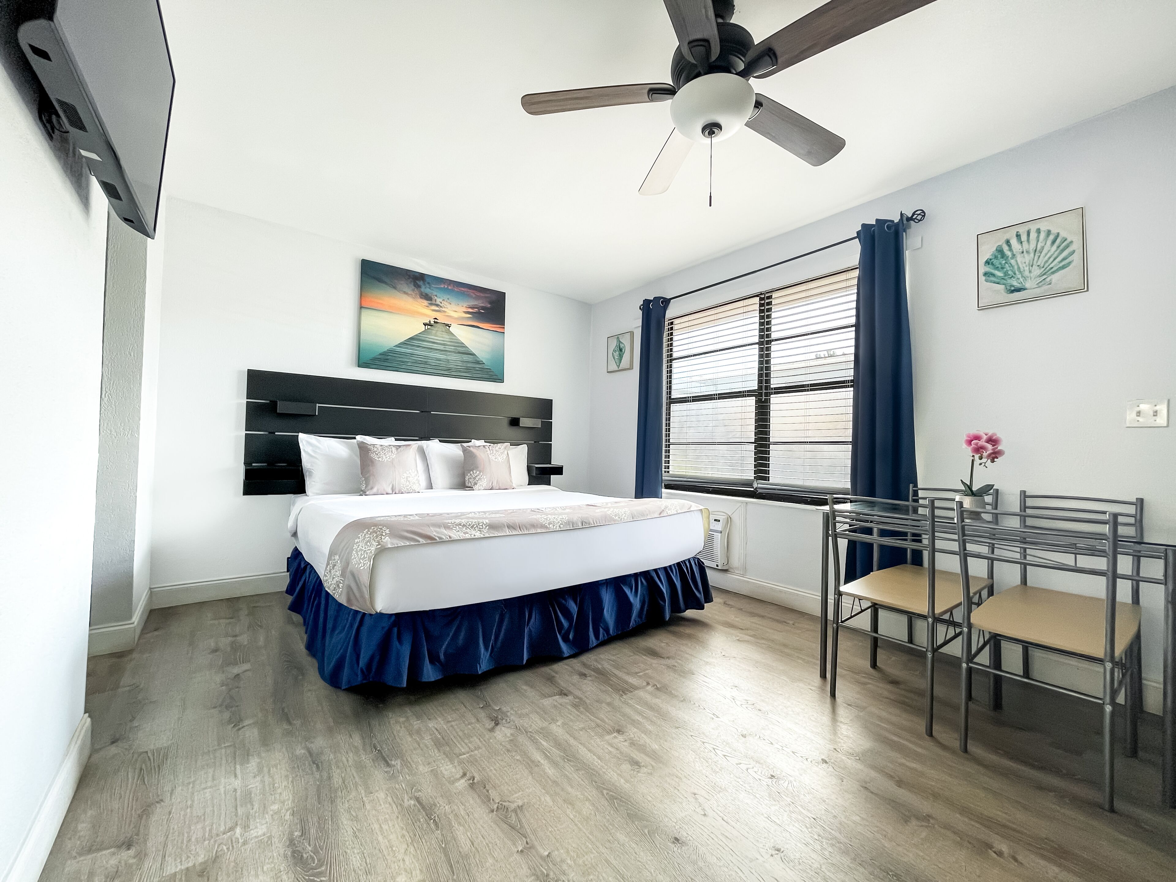 Une chambre à coucher avec un lit, un ventilateur de plafond, une fenêtre avec des stores, un bureau avec des chaises et un tableau accroché au mur.