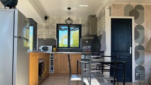 Una cocina moderna con refrigerador de acero inoxidable, armarios de madera, microondas y estufa a gas. Hay una ventana con vista a follaje, y un comedor con sillas metálicas y una mesa.