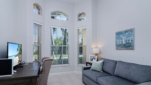A modern living room with a grey sofa, a wooden dining table, and a TV displaying a beach scene. Large windows with blinds allow natural light and views of greenery outside.