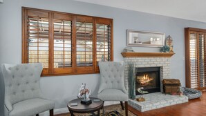 A living room with two grey upholstered chairs, a round coffee table, a fireplace, and large windows with wooden shutters.