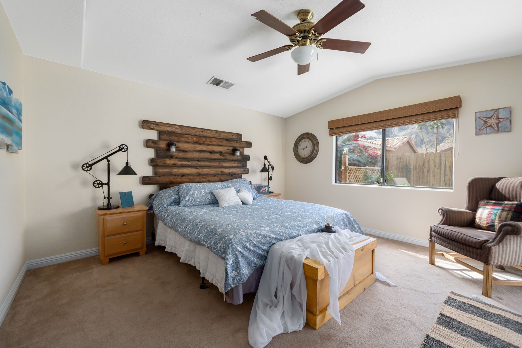 A bedroom with a bed, wooden headboard, nightstands, a chair, a ceiling fan, and a window with a view.