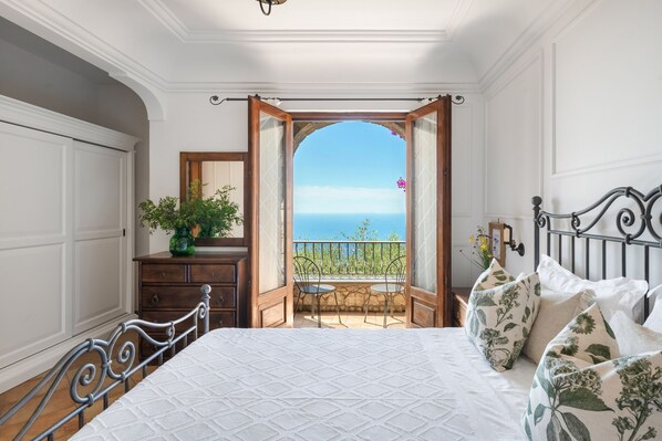 A bedroom with a bed, a dresser, a mirror, and a balcony with a view of the sea.