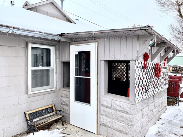 Un pequeño edificio de una sola planta con puerta y ventanas blancas, un banco en el exterior y una pared con celosía decorada con adornos rojos.