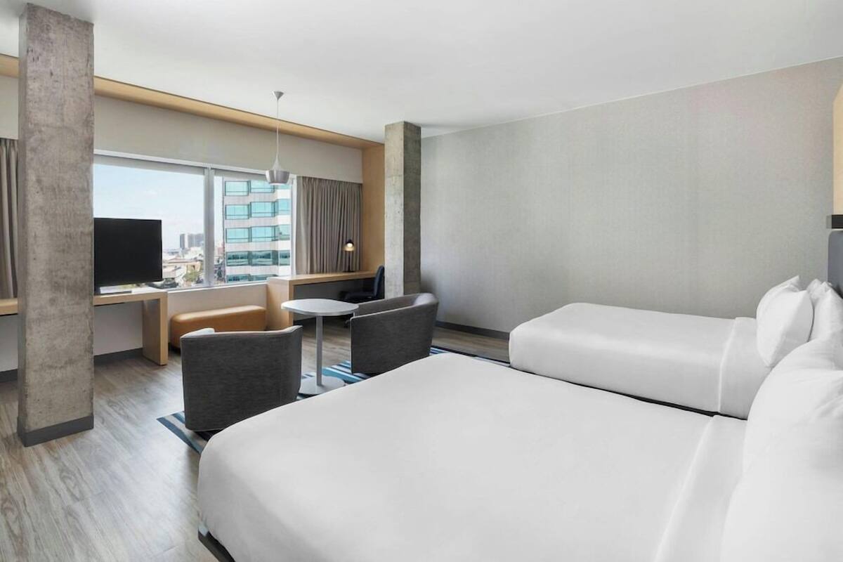 A hotel room with a bed, a desk with a chair, a small table, and a TV.