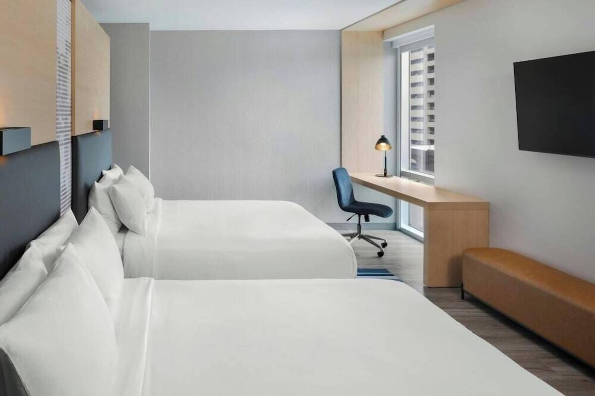 A hotel room with two beds, a desk, a chair, a bench, a TV, and a window with blinds.
