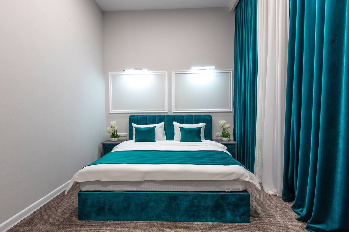 Quarto bem arrumado com cama, mesas de cabeceira e uma janela grande com cortinas.