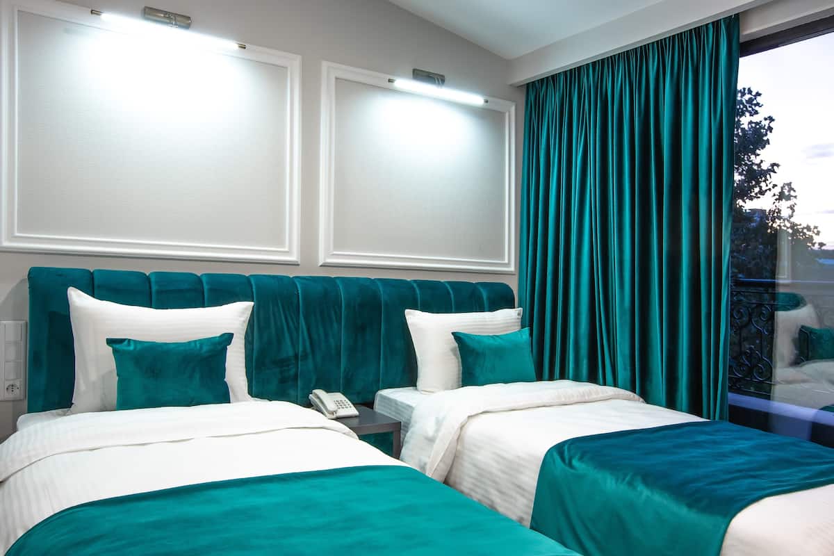 Quarto de hotel com duas camas, roupa de cama verde-água, cabeceira branca e uma janela grande com cortinas verde-água.