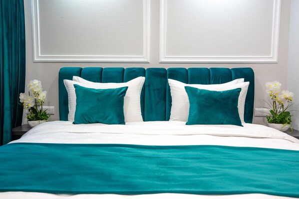 A neatly made bed with teal and white bedding, flanked by white pillows, in a room with a teal upholstered headboard and white walls. - Royal Inn (Tbilisi)