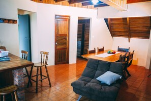 Un comedor con mesa y sillas de madera, un sofá negro con un cojín y piso de baldosas.