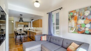 Een moderne woonkamer met een grijze modulaire bank, een eethoek met een tafel en stoelen, een keuken met houten kastjes en een kleurrijk abstract schilderij aan de muur.