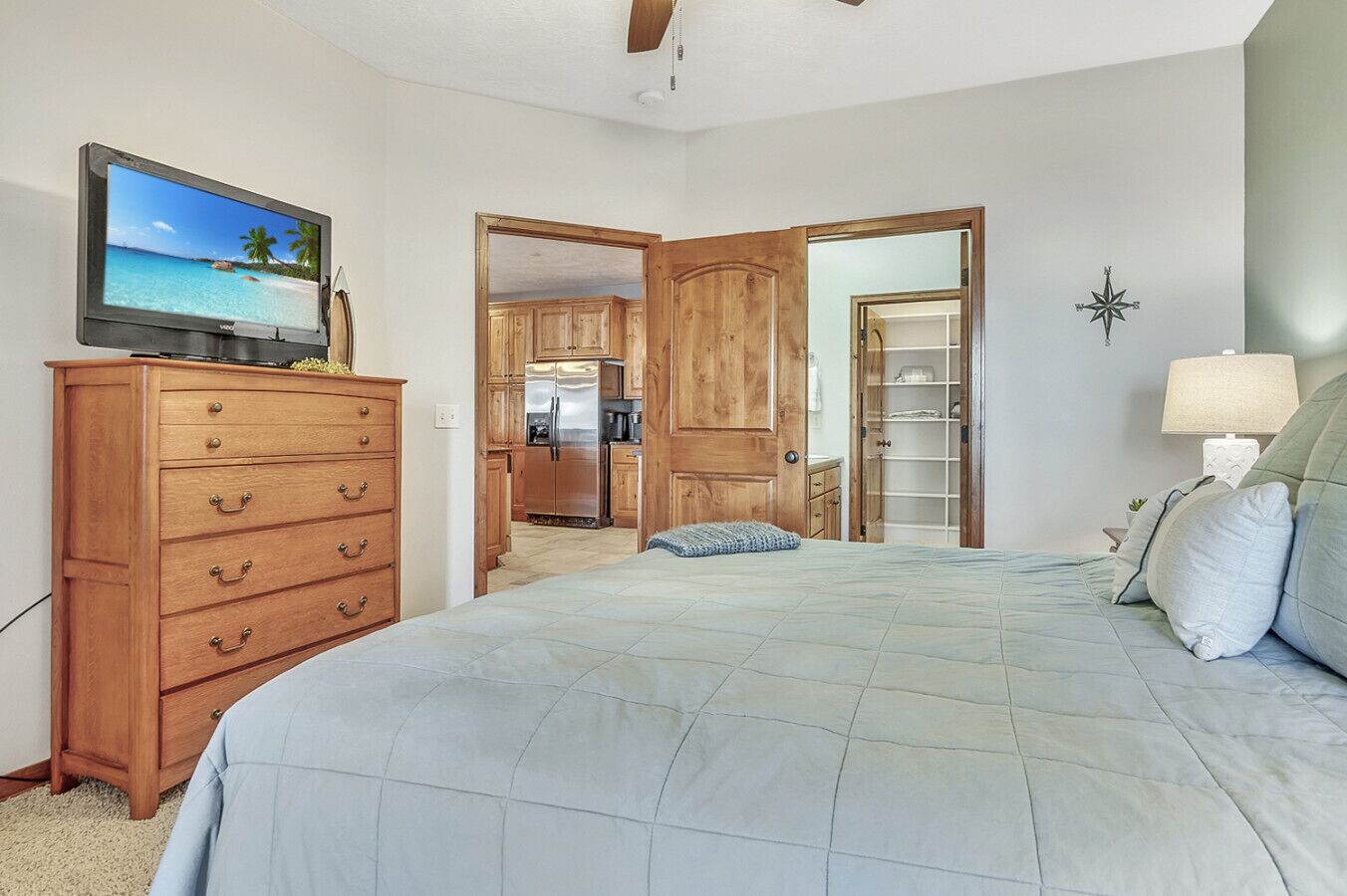 Une chambre à coucher avec un lit, une commode en bois, un téléviseur et un coin cuisine visible à travers une porte ouverte.