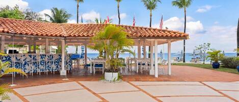 A covered outdoor seating area with a tiled roof, palm trees, and flags.