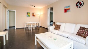 A modern living room with a white sofa, a dining table with chairs, and a coffee table. There is a painting on the wall and a clock. - Gabriel 01 by Interhome (Calpe/Calp)