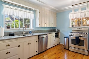 A kitchen with white cabinets, a stainless steel dishwasher, a stove, and a window with a view of trees. - Victorian Charm + BBQ Yard | Paso Robles Wine Country | Vacations on Vine (Paso Robles)