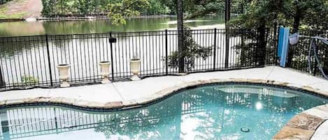 A swimming pool with a curved edge, surrounded by a stone deck and a metal fence.