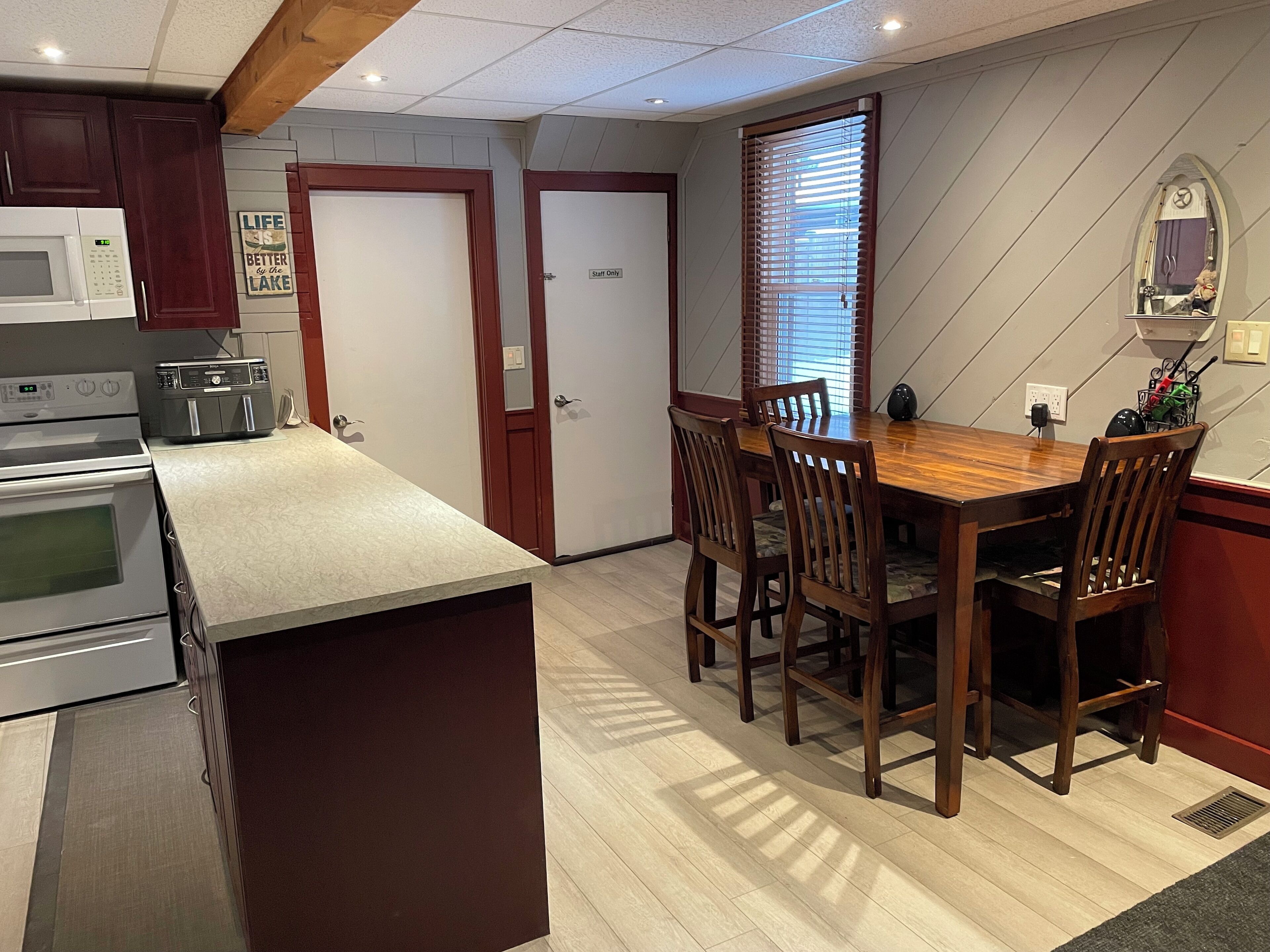 Une cuisine équipée d’une table et de chaises en bois, d’un four à micro-ondes et d’un grille-pain. Il y a deux portes : l’une est marquée « Réservé au personnel » et l’autre porte une pancarte indiquant : « La vie est plus belle au bord du lac. »