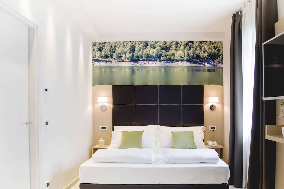 A hotel room with a bed, bedside lamps, a nightstand, a wall-mounted TV, and a large window with a forest view.