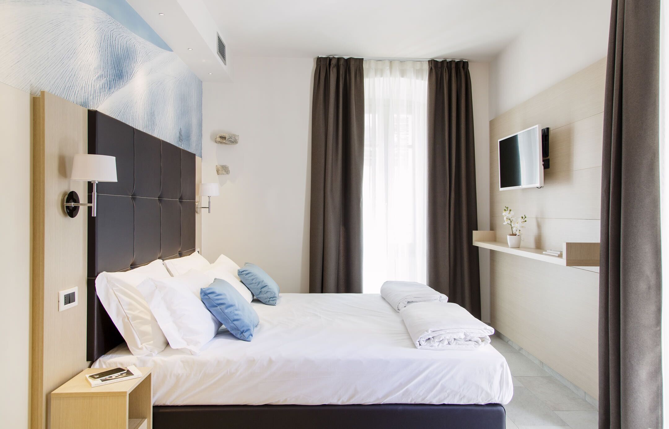 A modern hotel room with a large bed, a flat-screen TV, a bedside table, and a wall-mounted shelf with a vase.
