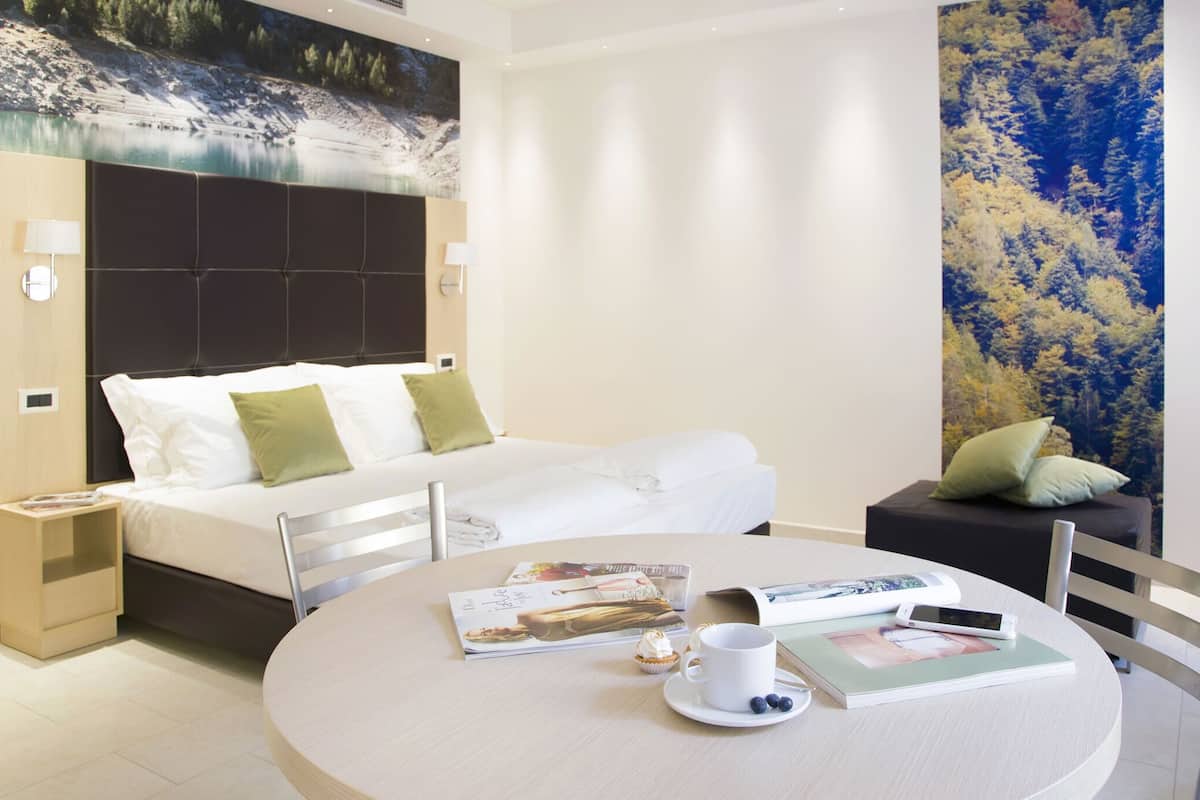 A modern hotel room with a bed, a round dining table with a magazine, a cup, and a smartphone.