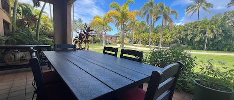 A covered patio with a table and chairs, overlooking a garden with palm trees and a lawn.
