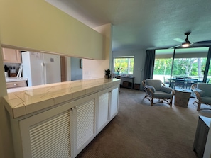A modern kitchen with a central island, a dining area with wicker furniture, and a view of greenery through large windows.