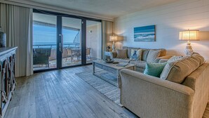 A spacious living room with a large sliding glass door leading to a balcony with a view of the ocean. There is a comfortable sofa set and a coffee table in the center of the room.