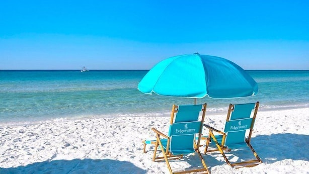 Two beach chairs with a turquoise umbrella on a sandy beach with clear blue water and a distant boat.