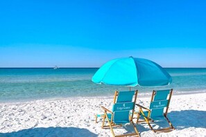 Two beach chairs with a turquoise umbrella on a sandy beach with clear blue water and a distant boat.